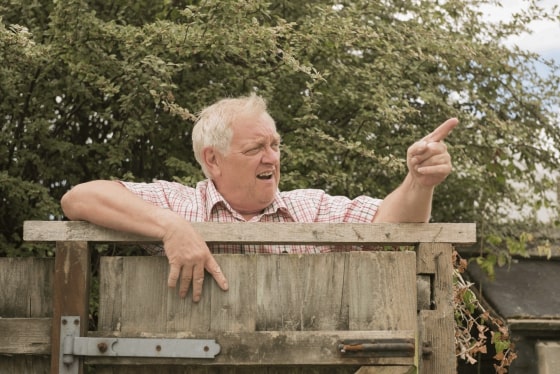 A old man pointing over his yard fence and looking annoyed